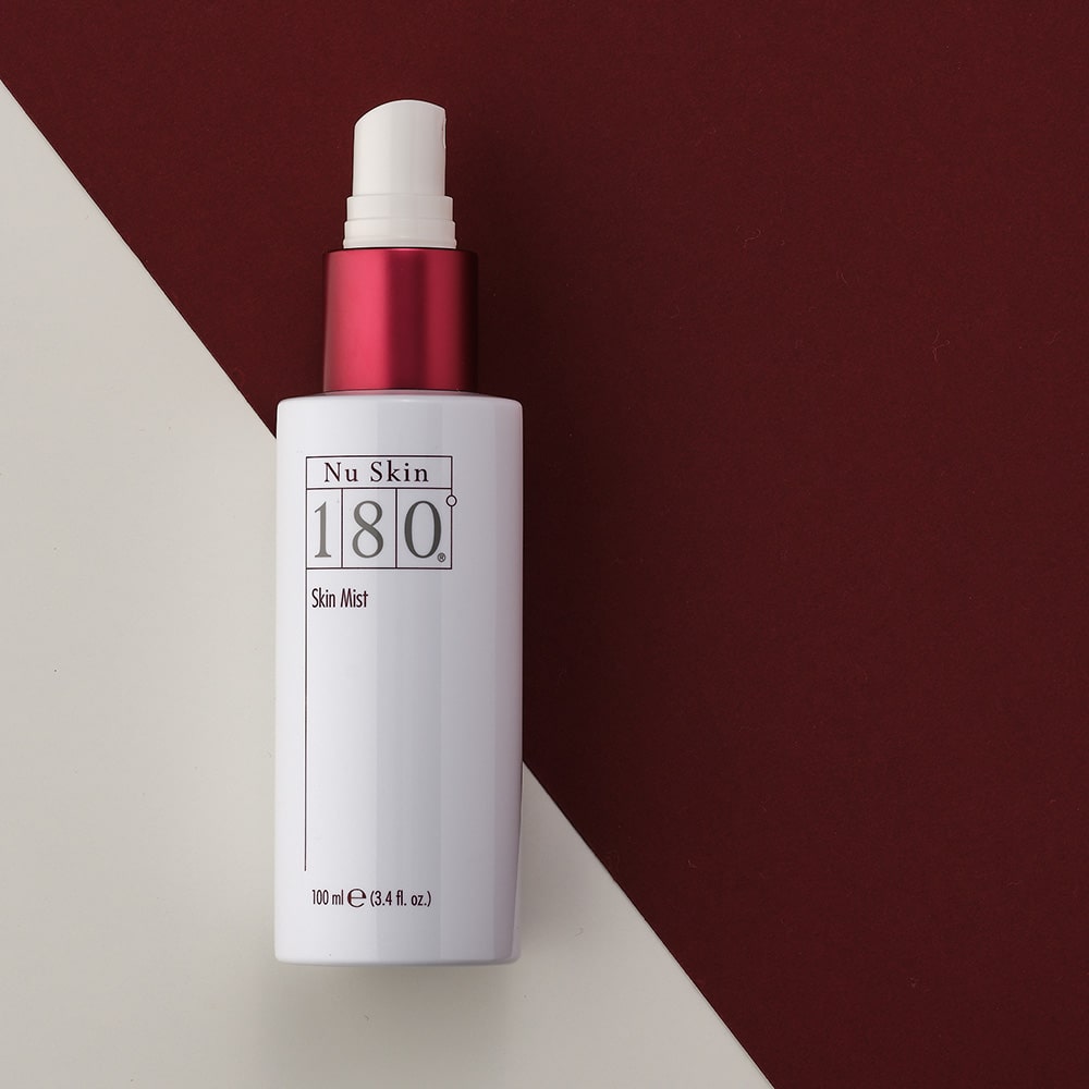 Gesichstwasser NuSkin 180° Skin Mist (100ml) liegt ohne Verschlusskappe auf weinroter und bescher Oberfläche.