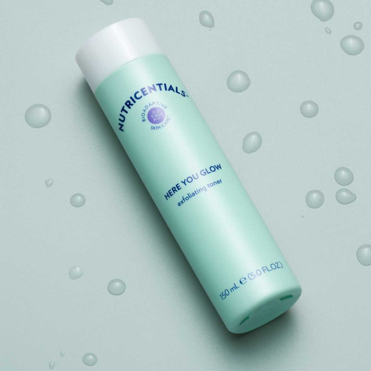 Nutricentials Here You Glow Exfoliating Toner, Tube (150ml) liegt geschlossen auf glatter hellgrüner Oberfläche. Die Tube ist von Wassertropfen umgeben.