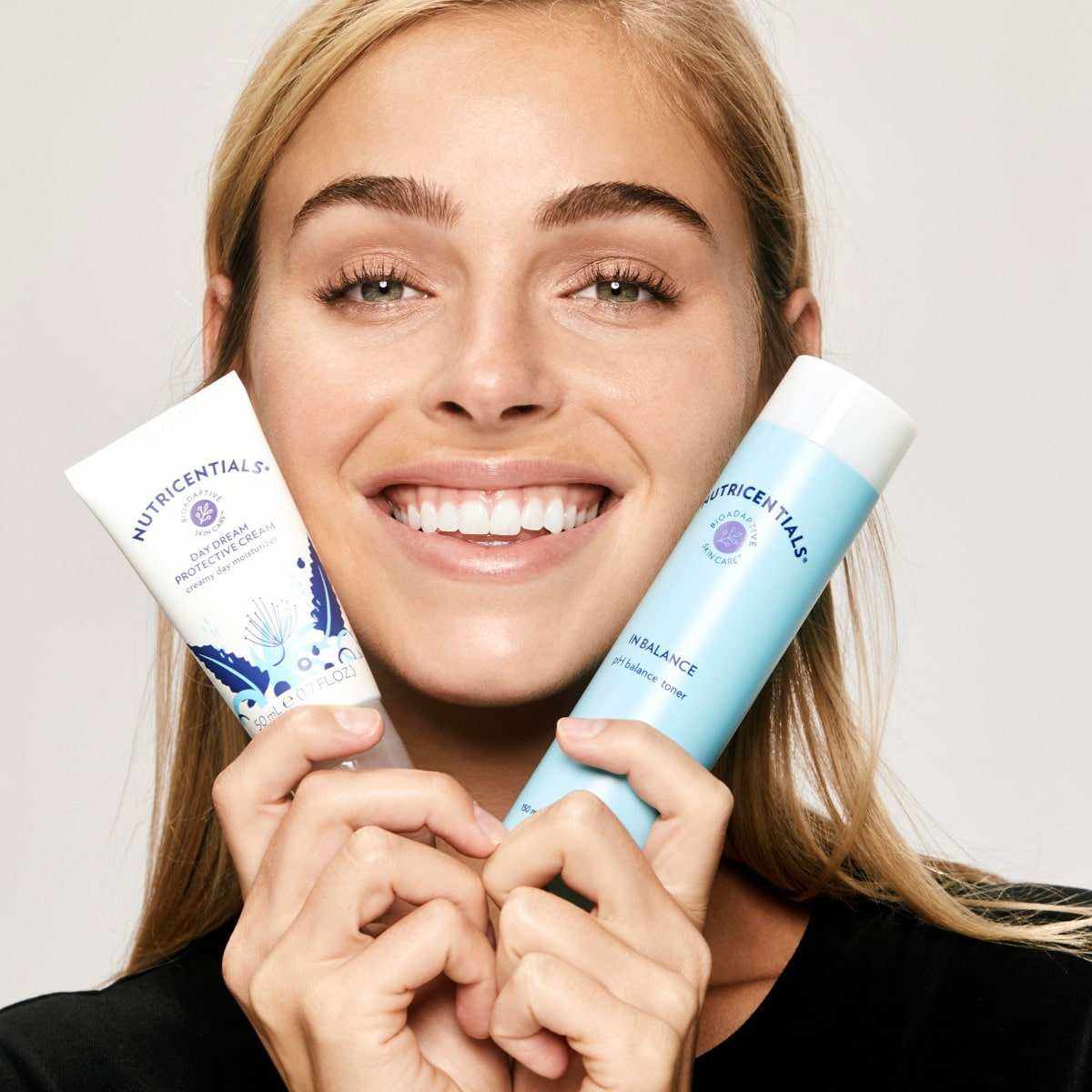 Junge Frau mit blonden Haaren und strahlendem Lächeln hält sich den In Balance pH Balance Toner und die Daydream Protective Cream (Nutricentials Produkte) an ihr Gesicht.