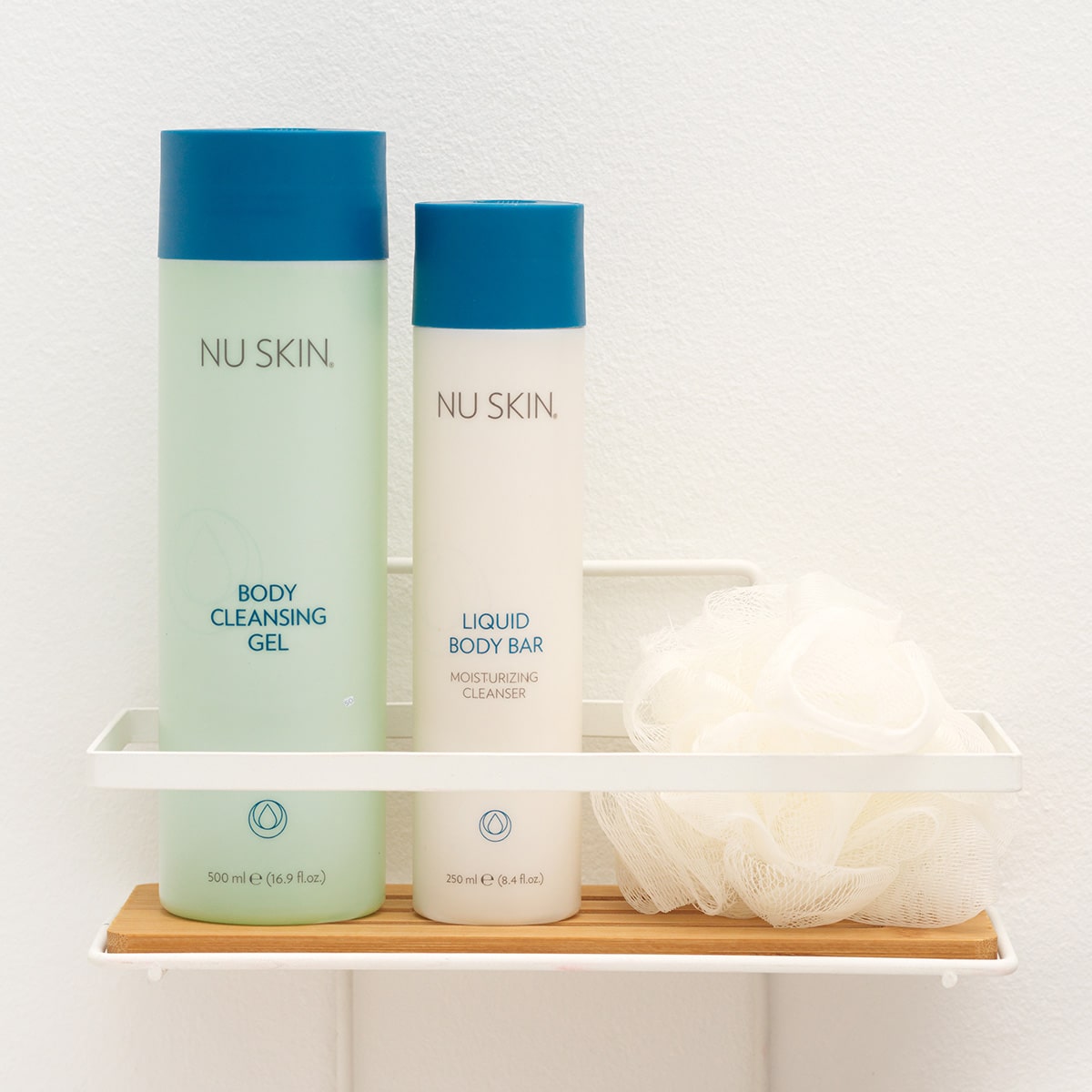 Nu Skin Basic Produkte (Body Cleansing Gel, 500ml und Liquid Body Bar, 250ml) stehen geschlossen auf einer kleinen Ablage in sanitärer Umgebung. Daneben liegt ein weißer Schwamm.