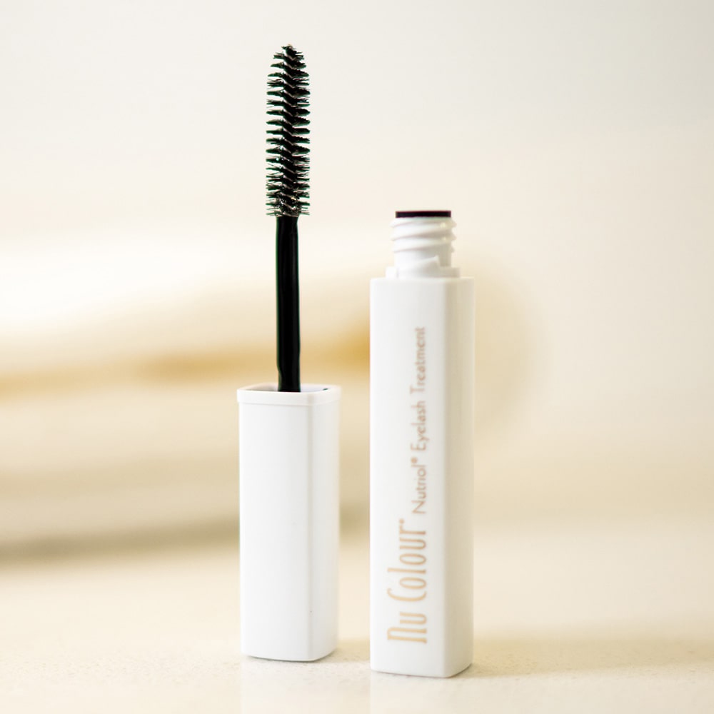 Produktfotografie. Front: Nu Colour Nutriol Eyelash Treatment von Nu Skin Tube (5ml) steht geöffnet in, von warmem Licht durchfluteter Umgebung.