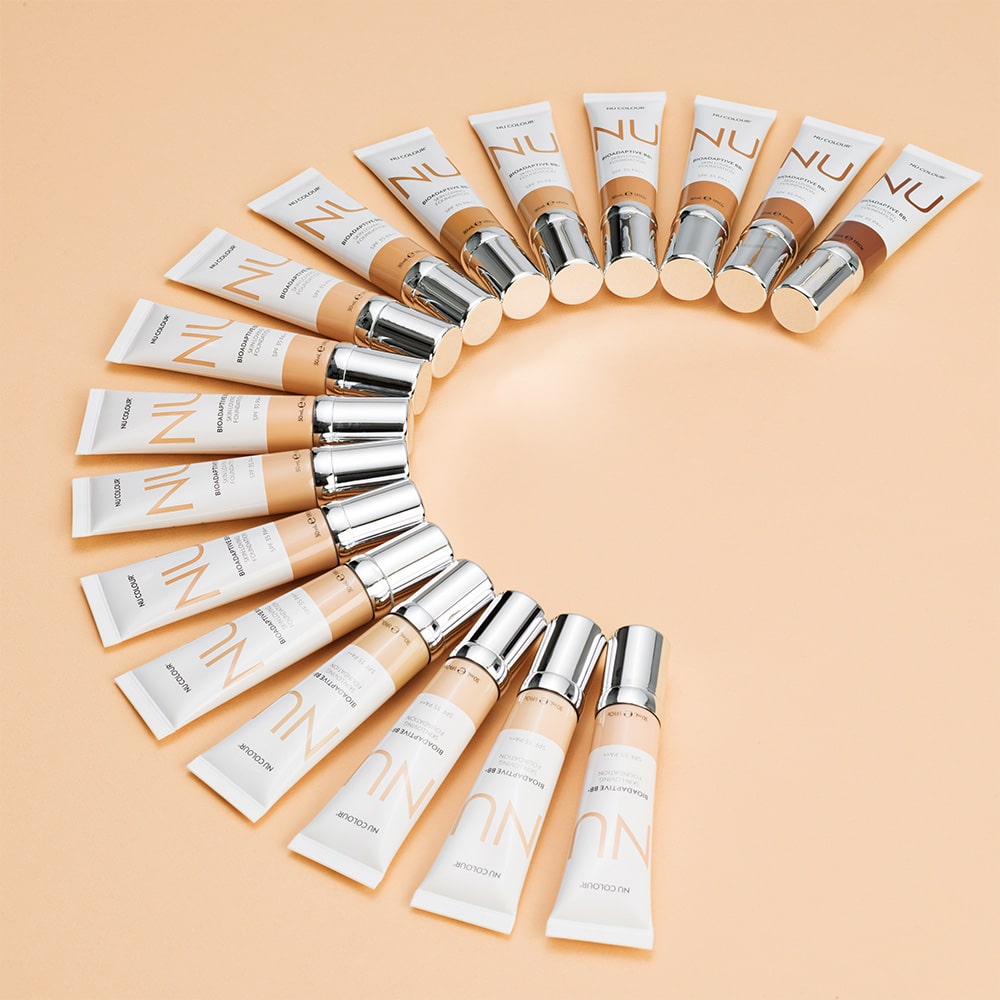 Nu Skin Nu Colour Bioadaptive BB+ Skin Loving Foundation Tuben (je 30ml) liegen halbkreisförmig, aufgefächert auf ockerfarbener Oberfläche.