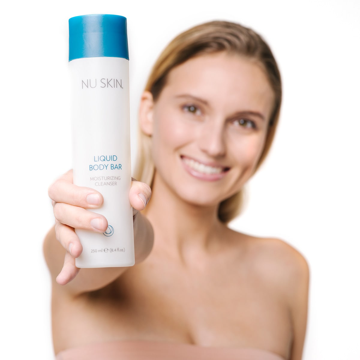 Junge Frau mit blonden nassen Haaren hält breit lächelnd Liquid Body Bar von Nu Skin (Tube, 250ml) vor sich in die Kamera.