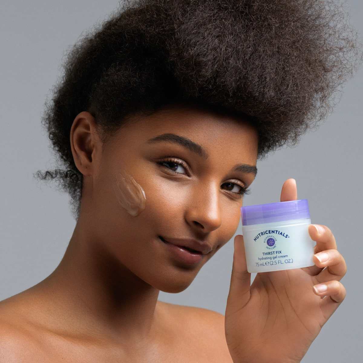 Junge Frau mit dunklem glatten Teint steht vor hellgrauem Hintergrund. Sie hält lächelnd Thirst Fix Hydrating Gel Cream von Nu Skin in der Hand. Auf ihrer Wange ist bereits eine kleine Menge des durchsichtigen Gels aufgetragen.